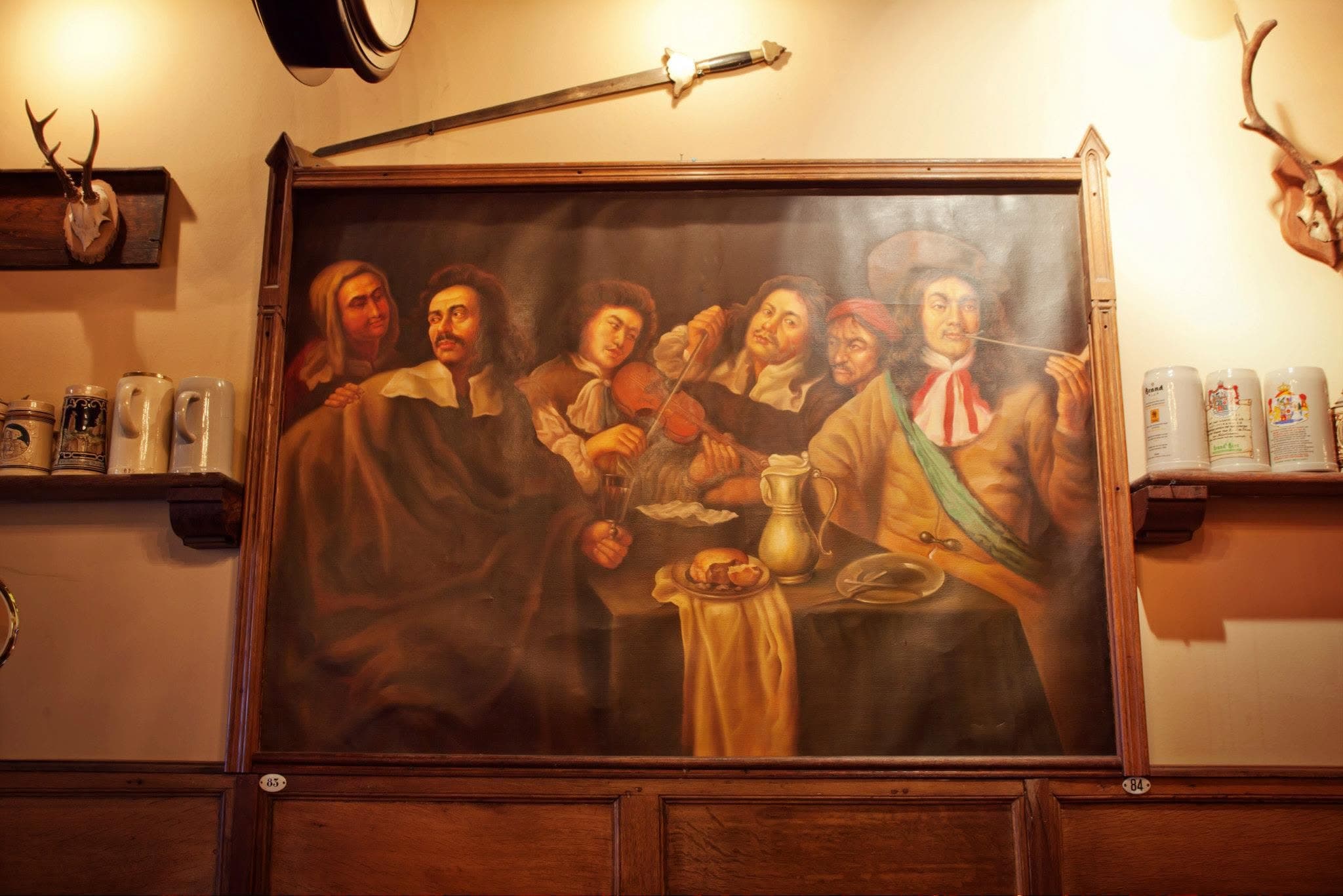 Klassiek schilderij van musicerend gezelschap aan de muur, tussen tinnen kroezen en geweien in het café